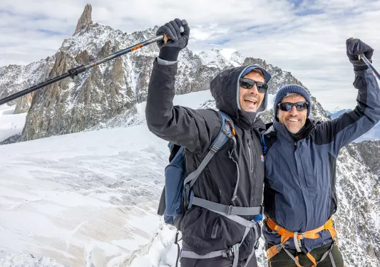 Two men in snow gear, holding up their arms while holding walking poles on top of a snowy mountain