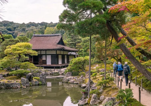 Three people walking on a small path on the right towards a Japanese-style building in the forest