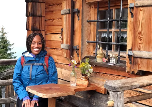 A person poses in front of a wooden house