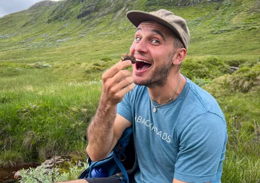 Man sitting in a grassy field with his mouth open eating a snack