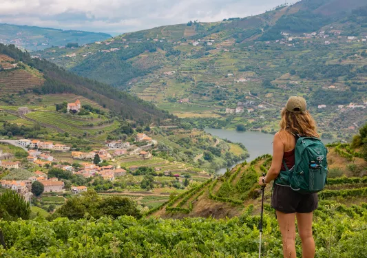 Woman wearing a backpack, standing on top of a hill looking down at a small town surrounded by trees