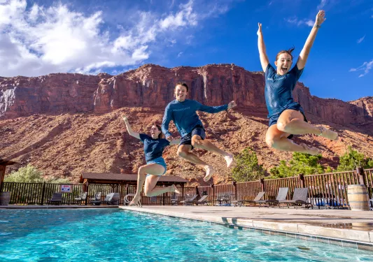 Three people jumping into an outdoor pool, with a canyon in the background