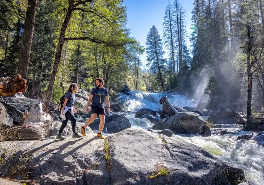 Two hikers cross rocks under a waterfall