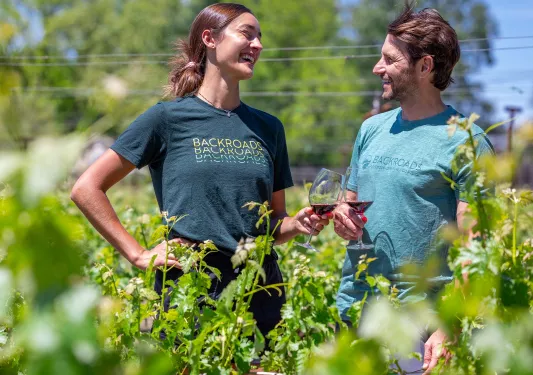 Man and woman smiling while holding up wine glasses, standing in a vineyard