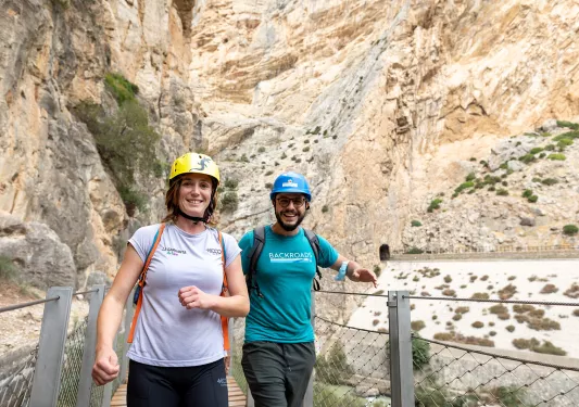 Man and woman walking on a wooden bridge with a large cliff in the background