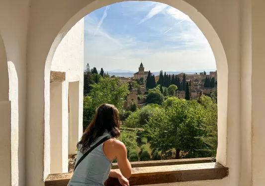 Woman standing next to a large window, looking out to a town surrounded by tall trees
