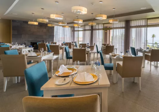 Restaurant dining hall with blue and beige cushioned chairs and large curtains along the windows