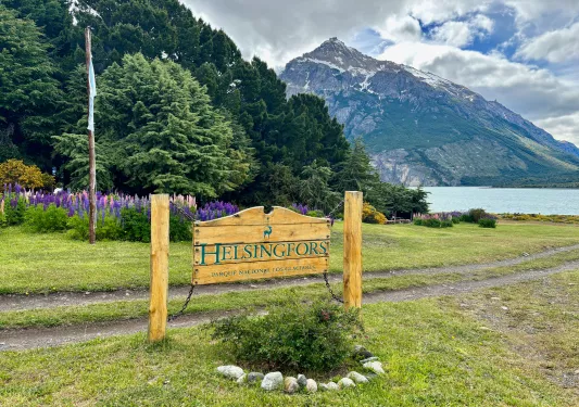 Wooden sign with the word "Helsingfors", in front of tall trees and an open lake
