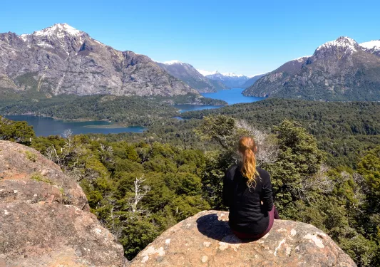 Woman sitting on a large boulder, looking to a valley of trees and a lake in the distance