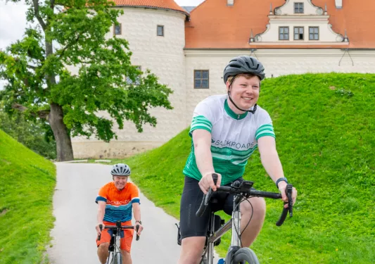 Two men smiling while riding bikes on a road, with a beige and orange castle-like building in the back