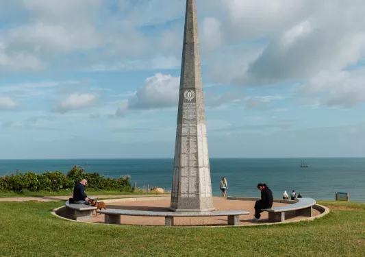 Pillar monument in the middle of a grass field, with the ocean in the background