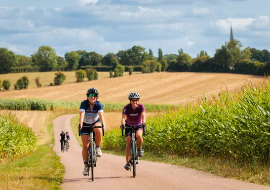 Two women riding bikes on a road, surrounded by fields of crops
