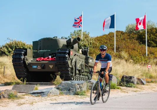 Man smiling while riding a bike, in front of a green tank and three flags
