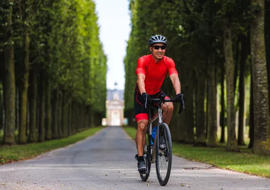 Man in red wearing sunglasses, riding their bike on a long row surrounded by trees