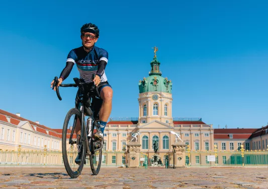 Man wearing biking gear, riding a bike away from a large palace-like building