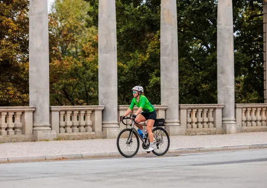 Woman wearing green biking gear, riding a bike on a road with large stone pillars in the background