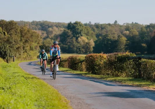 Two people smiling while riding their bikes on a road surrounded by bushes and trees