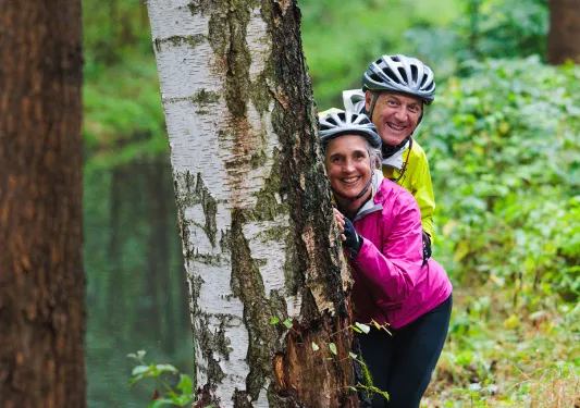 Man and woman wearing neon jackets, hiding behind a tree ina forest