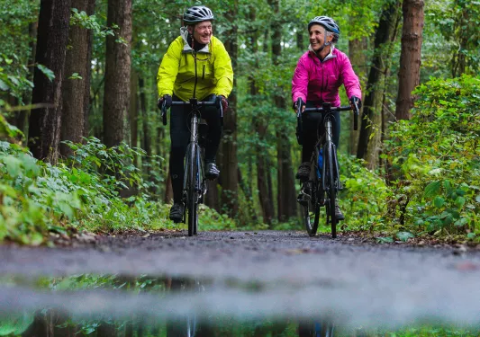 Man and woman in rain jackets riding bikes on a road, surrounded by tall trees