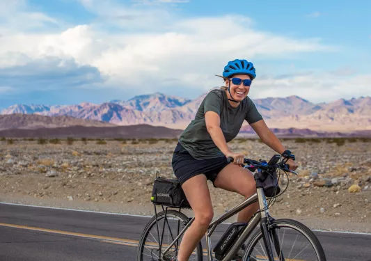 Woman smiling and riding a bike on a road, with mountains in the bckground
