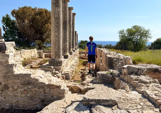 Man walking through temple ruins, with views of the ocean in the distance