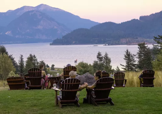 Groups of people sitting on wooden chairs outdoors, looking out to a large lake