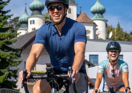 Man and woman smiling while biking, with a large palace in the backround