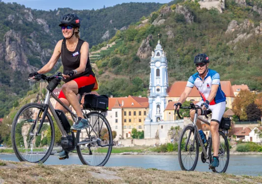 Man and woman biking on a dirt trail, with a river and palace building in the background