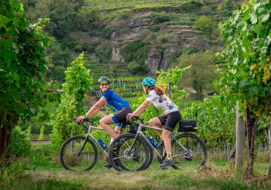 Two women looking at each other, smiling and biking through a vineyard