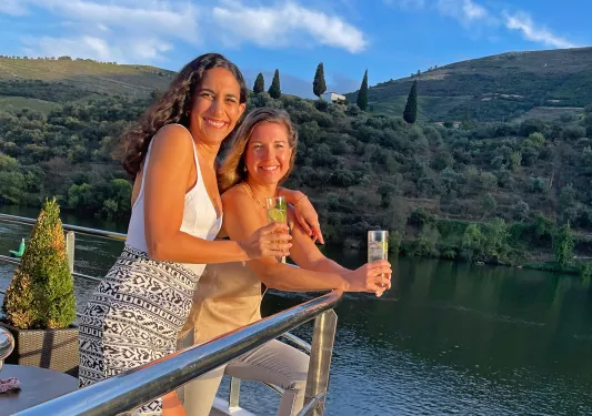 Two women smiling by a boat's railing, smiling while holding drinks