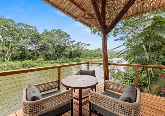 Outdoor balcony with two woven chairs and a circular table, with a river in the background
