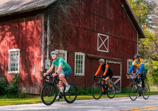 Three men riding bikes in front of a red barn
