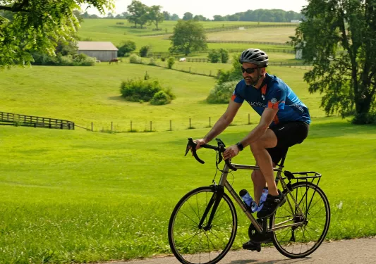 Man wearing biking gear and smiling while riding a bike on a road, with a large grass valley in the background