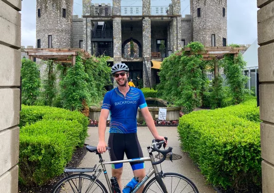 Man standing behind his bike, smiling with a large, stone building in the background
