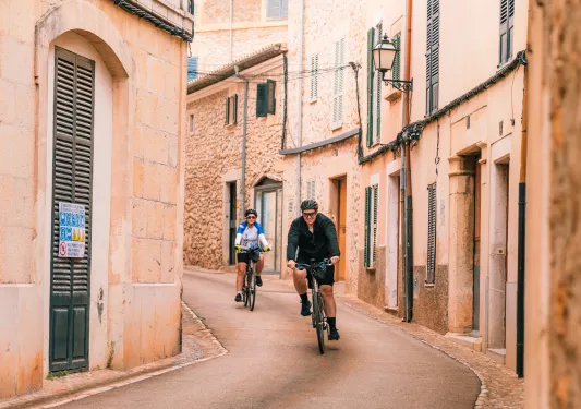Two people riding bikes along an alleyway in a town of brick buildings