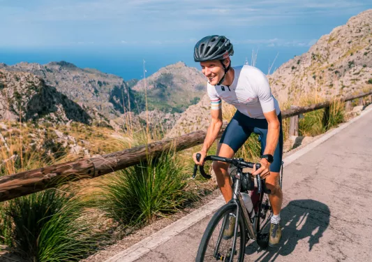 Man smiling while riding a bike on a road, with large cliffs in the distance