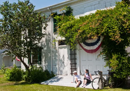 Two women sitting on a small flight of stairs in front of a white building, surrounded by trees and plants