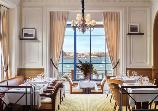 Restaurant dining hall with white chairs and tables, with large windows looking out to the ocean