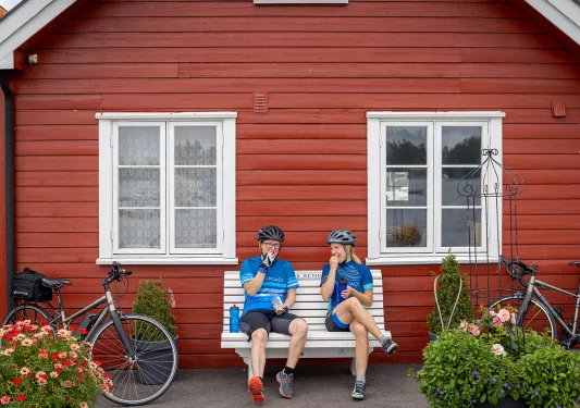 Man and woman sitting in front of a red house on a white bench, eating snacks