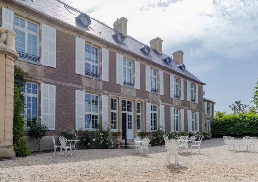 Exterior view of beige, stone building with a courtyard with white tables and chairs in front