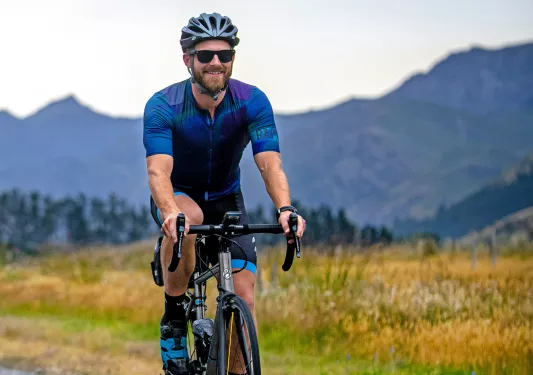 Man wearing biking gear, riding a bicycle on an asphalt road with tall mountains in the background