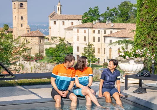 One man and two women sitting with their feet in a pool, with a rustic town in the background