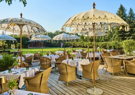 Outdoor patio with dining tables and woven chairs, with decorated umbrellas and a grass field in the background