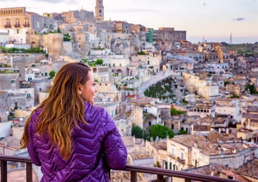 Woman wearing a purple jacket looking out of a balcony towards a town on a hill