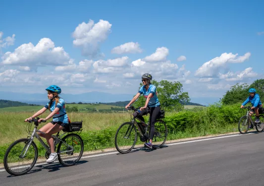 Woman and two kids riding their bikes on a road, with large grass valleys in the background