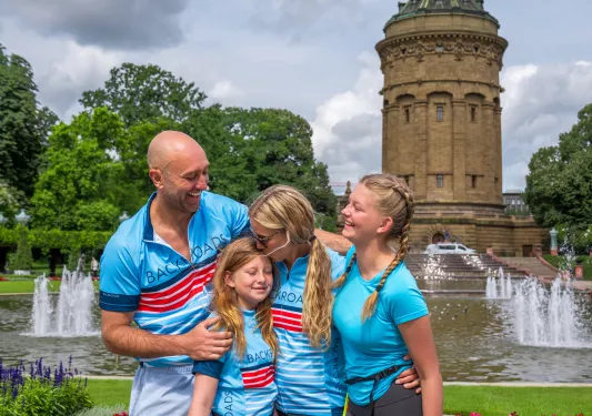 A family of bikers smiling and hugging, in front of a large fountain