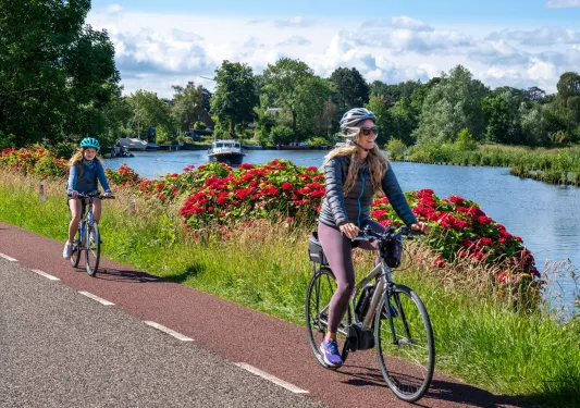 Woman and daughter riding their bike on a road, with flowers and a lake in the background