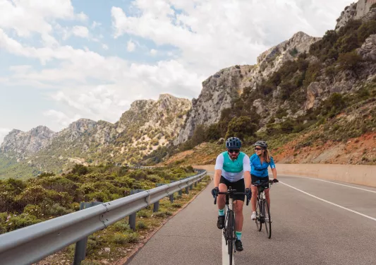 Man and woman riding bikes on an empty road, with large cliffs in the background