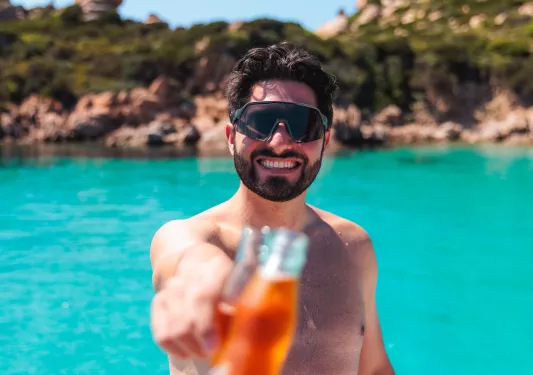 Man wearing sunglasses, raising a glass bottle with a lake in the background