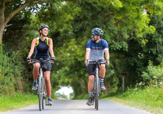 Man and woman smiling while riding their bikes on a road, with large trees surrounding